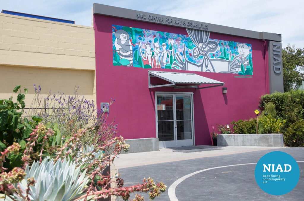 NIAD Art Center's facade: a magenta-colored building with a colorful mural and silver awning, with a planter full of succulents in the foreground