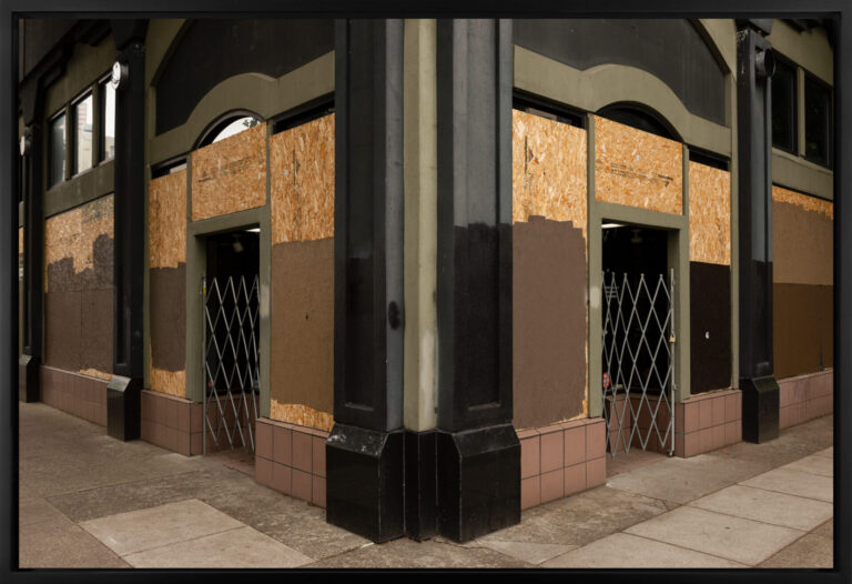 A color photograph depicts the corner of a commercial building. The windows surrounding two doorways have been covered with plywood, and much of the wood is painted brown